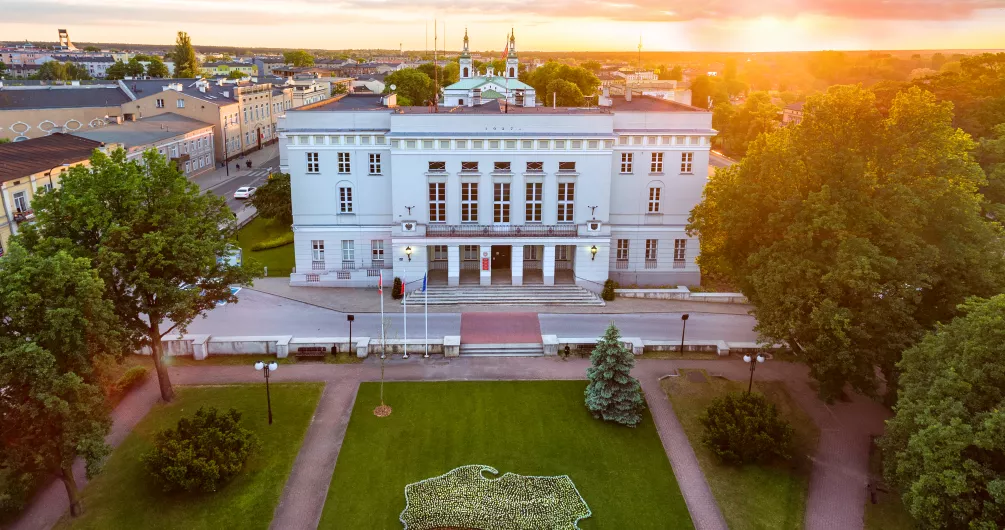 Zdjęcie przedstawia budynek Urzędu Miasta w Tomaszowie Mazowieckim podczas zachodu słońca. Przed urzędem skwer, na którym jest biało-czerwona flaga z kwiatów