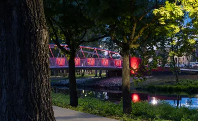 Zdjęcie przedstawia kładkę nad rzeką Wolbórką w Parku Bulwary. Oświetlona na czerwono